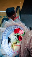 A bouquet featuring vibrant red and pink roses, a small teddy bear wearing a graduation cap, and decorative money arranged like flower petals is held by a person in a textured, light brown outfit. The background shows a person with a short hairstyle sitting in what appears to be an escalator setting.