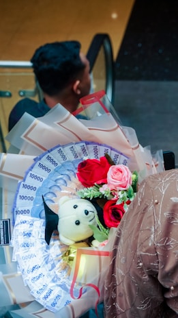 A bouquet featuring vibrant red and pink roses, a small teddy bear wearing a graduation cap, and decorative money arranged like flower petals is held by a person in a textured, light brown outfit. The background shows a person with a short hairstyle sitting in what appears to be an escalator setting.
