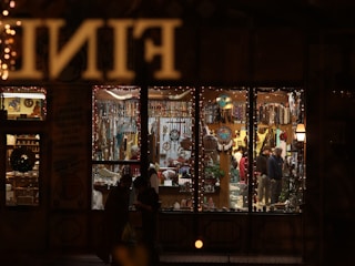 A warmly lit store is filled with various ornaments and trinkets, visible through large window panes. The interior is decorated with a variety of colorful items, including dreamcatchers and beads. People inside are browsing the shelves, while the exterior of the store is adorned with fairy lights, contributing to a festive atmosphere.
