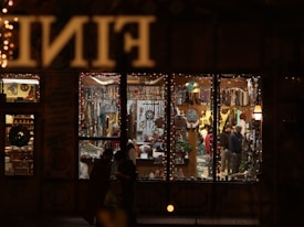 A warmly lit store is filled with various ornaments and trinkets, visible through large window panes. The interior is decorated with a variety of colorful items, including dreamcatchers and beads. People inside are browsing the shelves, while the exterior of the store is adorned with fairy lights, contributing to a festive atmosphere.