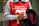 A person wearing a red apron with white text and holding a donation cup. The text mentions 'St-Vincent de Paul'. The individual also carries a coat and has a braided hairstyle.