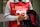 A person wearing a red apron with white text and holding a donation cup. The text mentions 'St-Vincent de Paul'. The individual also carries a coat and has a braided hairstyle.