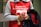 A person wearing a red apron with white text and holding a donation cup. The text mentions 'St-Vincent de Paul'. The individual also carries a coat and has a braided hairstyle.