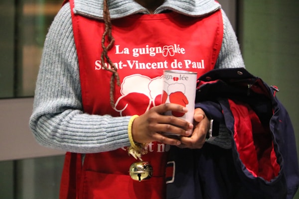 A person wearing a red apron with white text and holding a donation cup. The text mentions 'St-Vincent de Paul'. The individual also carries a coat and has a braided hairstyle.