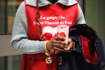 A person wearing a red apron with white text and holding a donation cup. The text mentions 'St-Vincent de Paul'. The individual also carries a coat and has a braided hairstyle.