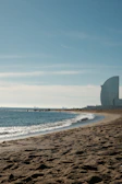 A serene beach with golden sand and gentle waves lapping against the shore. In the distance, a modern, sail-shaped building rises into the clear blue sky, while a few people are scattered along the shoreline. The horizon meets the sky in soft hues.