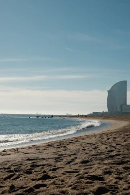 A serene beach with golden sand and gentle waves lapping against the shore. In the distance, a modern, sail-shaped building rises into the clear blue sky, while a few people are scattered along the shoreline. The horizon meets the sky in soft hues.