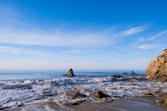A serene coastal shoreline with gentle waves washing over engineered rock barriers under a clear blue sky.