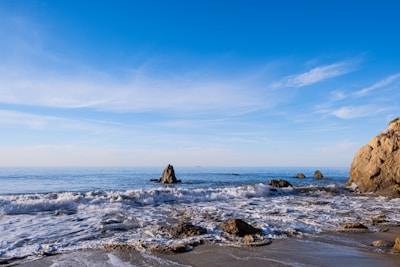 A serene coastal shoreline with gentle waves washing over engineered rock barriers under a clear blue sky.