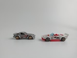 Two toy cars are placed side by side on a plain background. The car on the left is gray with red and black details, featuring a closed top design. The wheels have a bronze-colored rim. The car on the right is white with red accents and an open-top design with red seats, featuring a blue windshield and black wheels.