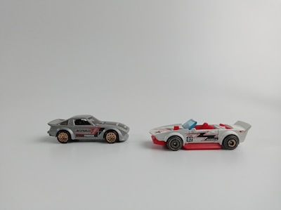 Two toy cars are placed side by side on a plain background. The car on the left is gray with red and black details, featuring a closed top design. The wheels have a bronze-colored rim. The car on the right is white with red accents and an open-top design with red seats, featuring a blue windshield and black wheels.