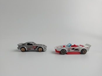 Two toy cars are placed side by side on a plain background. The car on the left is gray with red and black details, featuring a closed top design. The wheels have a bronze-colored rim. The car on the right is white with red accents and an open-top design with red seats, featuring a blue windshield and black wheels.
