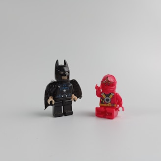 Two LEGO mini-figures are positioned against a plain background. One is dressed as a superhero in a black costume and mask, while the other is in a red ninja outfit with detailed armor.