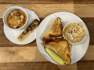 A wooden table holds two plates of food. The first plate contains a bowl of tomato soup topped with croutons and cheese, accompanied by a garlic breadstick. The second plate features a grilled sandwich with a side of coleslaw in a small dish and a pickle spear.