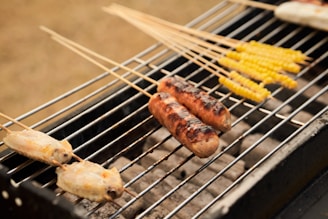 Grilled food items such as sausages, corn, and chicken are placed on skewers over an outdoor barbecue grill. The sausages display a charred texture, while the corn appears yellow and grilled. The skewer setup indicates a casual outdoor cooking scene.