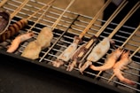 A variety of skewered seafood and meats are grilling on a barbecue. The skewers are laid out in a row, with pieces of shrimp, squid, and other meats visible, all lightly seasoned.