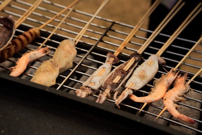 A variety of skewered seafood and meats are grilling on a barbecue. The skewers are laid out in a row, with pieces of shrimp, squid, and other meats visible, all lightly seasoned.
