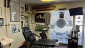A tattoo studio is filled with various artworks and decorations. The walls display multiple framed pictures and posters, some with intricate designs and others featuring artistic portraits. There is a tattoo chair positioned in front of the displayed artworks. A surfboard is mounted above, with a hat hanging from it. Nearby shelves and surfaces hold tattooing equipment and various bottles, likely inks. The room has a clean and organized appearance, with a TV mounted in the corner and a large window with blinds, through which a parking lot can be seen.