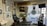 A tattoo studio is filled with various artworks and decorations. The walls display multiple framed pictures and posters, some with intricate designs and others featuring artistic portraits. There is a tattoo chair positioned in front of the displayed artworks. A surfboard is mounted above, with a hat hanging from it. Nearby shelves and surfaces hold tattooing equipment and various bottles, likely inks. The room has a clean and organized appearance, with a TV mounted in the corner and a large window with blinds, through which a parking lot can be seen.