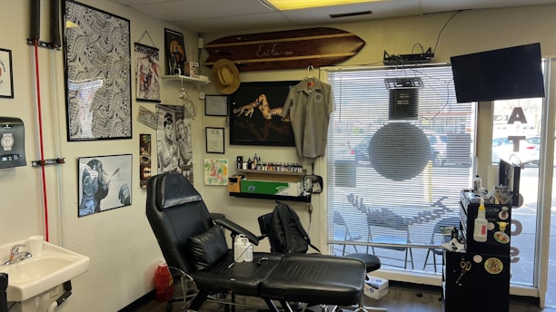 A tattoo studio is filled with various artworks and decorations. The walls display multiple framed pictures and posters, some with intricate designs and others featuring artistic portraits. There is a tattoo chair positioned in front of the displayed artworks. A surfboard is mounted above, with a hat hanging from it. Nearby shelves and surfaces hold tattooing equipment and various bottles, likely inks. The room has a clean and organized appearance, with a TV mounted in the corner and a large window with blinds, through which a parking lot can be seen.