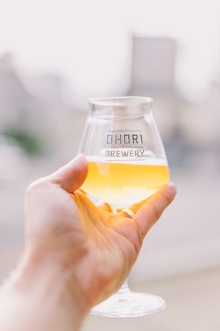 A hand is holding a clear glass filled with a light amber beverage, featuring the text 'OHORI BREWERY'. The background is blurred, suggesting a serene outdoor setting.