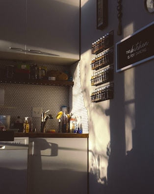 A cozy kitchen corner bathed in warm, late-afternoon sunlight. The wall is decorated with a hexagonal tile backsplash and shelves holding rows of spice jars. A sign reads 'This kitchen is for dancing'. Shadows of nearby objects are cast on the wall, adding artistic flair to the space.