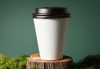 A white paper coffee cup with a dark brown plastic lid is placed on a tree stump, surrounded by green moss. The background is a muted green color, providing a natural setting for the scene.