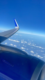 An airplane wing and engine are visible against a backdrop of scattered clouds and a vast, clear blue sky stretching to the horizon.