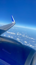 An airplane wing and engine are visible against a backdrop of scattered clouds and a vast, clear blue sky stretching to the horizon.