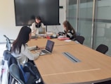 Three individuals are seated around a conference table in a meeting room, using laptops and focusing on their tasks. A large screen is mounted on the wall, and various office supplies are visible on the table. The room is well-lit, with modern office chairs and a glass partition visible in the background.