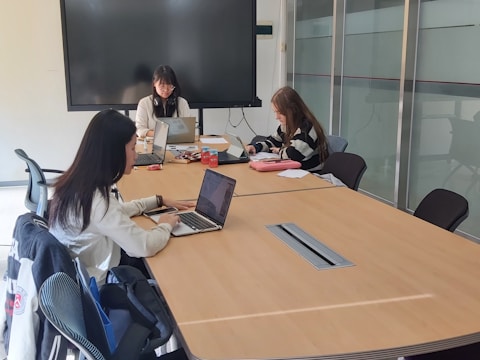 Three individuals are seated around a conference table in a meeting room, using laptops and focusing on their tasks. A large screen is mounted on the wall, and various office supplies are visible on the table. The room is well-lit, with modern office chairs and a glass partition visible in the background.