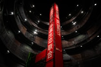 A tall, red vertical banner with text in both English and another language stands in the center of a large, circular multi-level structure. The structure is dimly lit, with lights highlighting the industrial design. A person is visible walking on one of the upper levels.