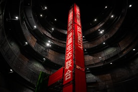 A tall, red vertical banner with text in both English and another language stands in the center of a large, circular multi-level structure. The structure is dimly lit, with lights highlighting the industrial design. A person is visible walking on one of the upper levels.