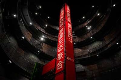 A tall, red vertical banner with text in both English and another language stands in the center of a large, circular multi-level structure. The structure is dimly lit, with lights highlighting the industrial design. A person is visible walking on one of the upper levels.