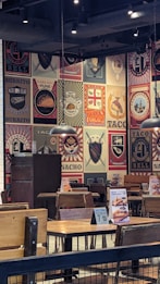 The image depicts the interior of a restaurant with walls covered in colorful and artistic vintage-style logos and advertisements for tacos and burritos. Wooden tables and chairs are arranged neatly, and the lighting is provided by overhead lamps, creating a cozy atmosphere.