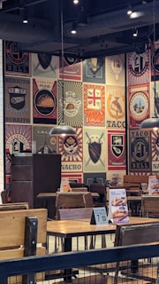 The image depicts the interior of a restaurant with walls covered in colorful and artistic vintage-style logos and advertisements for tacos and burritos. Wooden tables and chairs are arranged neatly, and the lighting is provided by overhead lamps, creating a cozy atmosphere.