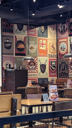 The image depicts the interior of a restaurant with walls covered in colorful and artistic vintage-style logos and advertisements for tacos and burritos. Wooden tables and chairs are arranged neatly, and the lighting is provided by overhead lamps, creating a cozy atmosphere.