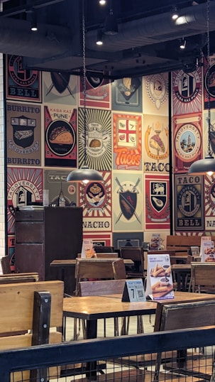 The image depicts the interior of a restaurant with walls covered in colorful and artistic vintage-style logos and advertisements for tacos and burritos. Wooden tables and chairs are arranged neatly, and the lighting is provided by overhead lamps, creating a cozy atmosphere.