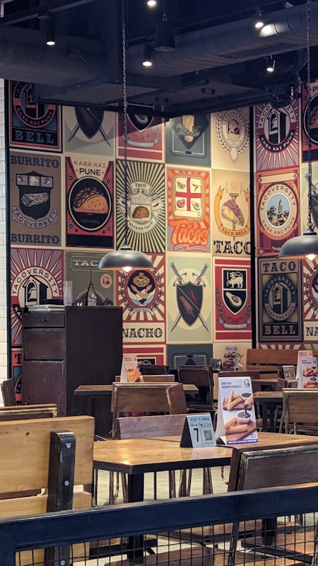 The image depicts the interior of a restaurant with walls covered in colorful and artistic vintage-style logos and advertisements for tacos and burritos. Wooden tables and chairs are arranged neatly, and the lighting is provided by overhead lamps, creating a cozy atmosphere.