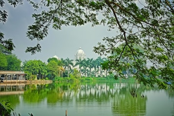 A serene landscape featuring a calm body of water with a reflection of surrounding trees and a white domed building in the background. The foreground includes leafy branches framing the image, adding depth and a natural feel. A small structure labeled 'Tripura Swimming Club' is visible on the left.