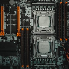A detailed view of a computer motherboard featuring two CPU sockets, various slots for RAM, and other components. The layout includes orange slots and connectors that add contrast to the predominantly gray and black color scheme, with intricate patterns of circuits visible throughout.