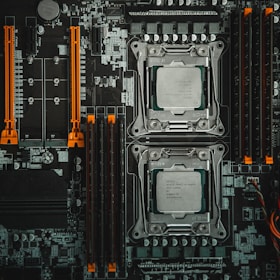 A detailed view of a computer motherboard featuring two CPU sockets, various slots for RAM, and other components. The layout includes orange slots and connectors that add contrast to the predominantly gray and black color scheme, with intricate patterns of circuits visible throughout.