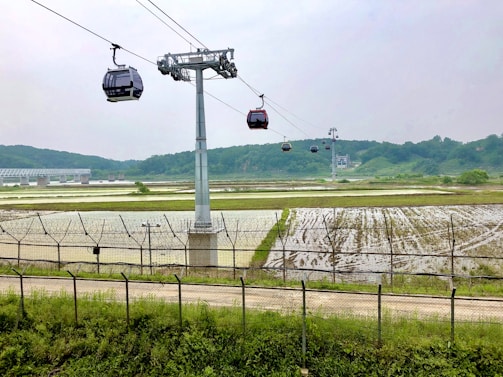 Cable cars are suspended from a metal cable over an expansive landscape featuring fields and a clear sky. In the distance, there is a bridge crossing a river with lush green hills beyond. A metal fence runs parallel to a dirt road at the base of the scene.