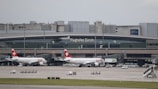 Two Swiss Air airplanes are parked at a terminal at Zurich Airport. The terminal building is large and modern with signage displaying 'Flughafen Zürich'. Various airport vehicles and equipment are scattered around the tarmac.