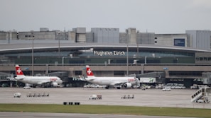 Two Swiss Air airplanes are parked at a terminal at Zurich Airport. The terminal building is large and modern with signage displaying 'Flughafen Zürich'. Various airport vehicles and equipment are scattered around the tarmac.