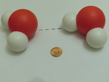 A model of two water molecules is depicted, with each consisting of one large red sphere representing oxygen and two smaller white spheres representing hydrogen. They are placed with a dotted line between them, suggesting a hydrogen bond. A coin is positioned below the molecules for scale reference.