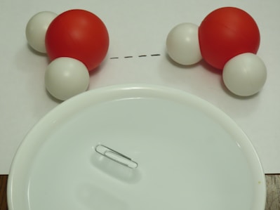 Two molecular models, each consisting of one large red sphere and two smaller white spheres, are positioned facing each other on a white surface. A white plate and a paper clip are placed in the foreground.