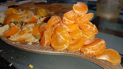 A pile of peeled tangerine segments stacked neatly on a wooden board. In the background, there are scattered tangerine peels. The setup appears casual, with the focus on the bright, juicy segments in the foreground.