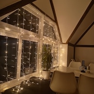 A cozy indoor scene featuring a large window adorned with string lights, casting a warm glow. There is a potted plant placed near the window. The room includes a white dining table with matching chairs, and a decorative vase on the table.