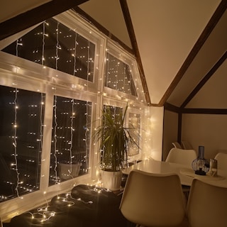A cozy indoor scene featuring a large window adorned with string lights, casting a warm glow. There is a potted plant placed near the window. The room includes a white dining table with matching chairs, and a decorative vase on the table.